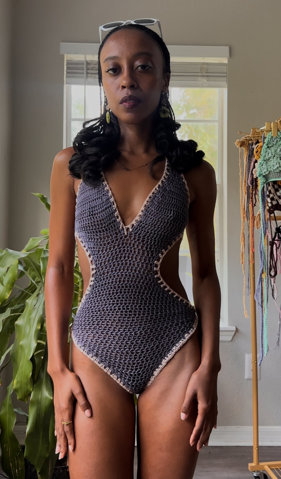 Woman wearing a patterned swimsuit indoors with a plant and clothing rack in the background.